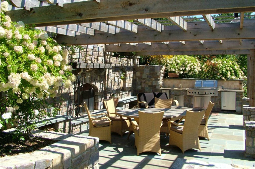 A landing formed by a retaining wall was used to create a combination kitchen/living room area away from it all. In addition to the grill and dining area, the space includes a fireplace and lounging furniture, capped by a pergola that would accommodate climbing foliage. This more remote space, also designed by Hess Landscape Architects, includes amenities such as a refrigerator and sink.